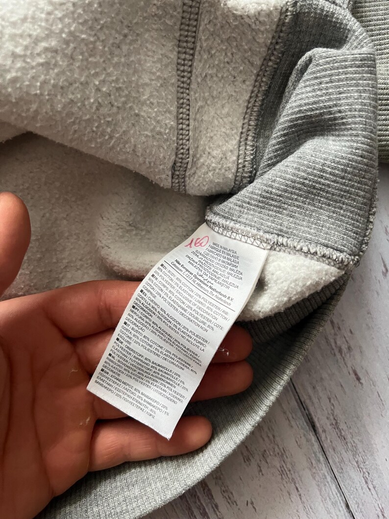 Puede incluir: Primer plano del interior de una sudadera gris con un forro blanco y difuso. Una etiqueta con texto en varios idiomas es visible. El exterior de la sudadera es de tejido de punto acanalado. Se ve la mano que sostiene la sudadera.