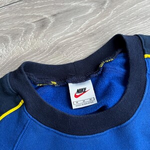 May include: Blue Nike sweatshirt with a navy blue collar and yellow accents. The Nike logo is visible on a white tag. The sweatshirt is a medium size and made in Malaysia.