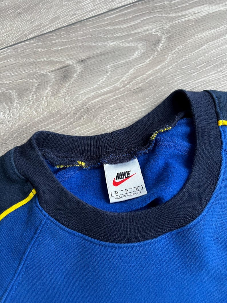 May include: Blue Nike sweatshirt with a navy blue collar and yellow accents. The Nike logo is visible on a white tag. The sweatshirt is a medium size and made in Malaysia.