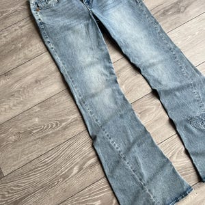 May include: Light blue denim jeans featuring a flared leg silhouette, a mid-rise waist with a button and zipper fly closure, five pockets, and a frayed raw edge waistband. The fabric has a faded wash finish with subtle whiskering at the hips.