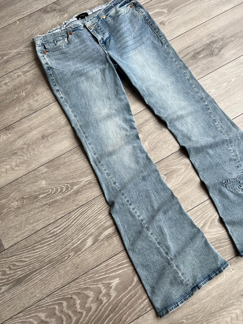 May include: Light blue denim jeans featuring a flared leg silhouette, a mid-rise waist with a button and zipper fly closure, five pockets, and a frayed raw edge waistband. The fabric has a faded wash finish with subtle whiskering at the hips.