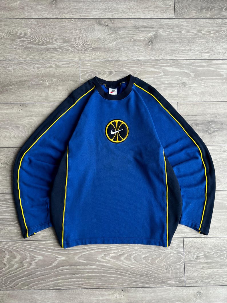 May include: Blue long-sleeve sweatshirt with a basketball graphic. The sweatshirt has black sleeves with yellow trim and a black collar. A yellow and black basketball logo is centered on the chest. The Nike logo is on the collar.
