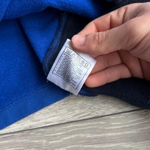 May include: Close-up of a blue and navy blue fleece garment. A white tag with care instructions is visible. The tag includes symbols for washing, ironing, and other care details. The garment appears to be soft and warm.