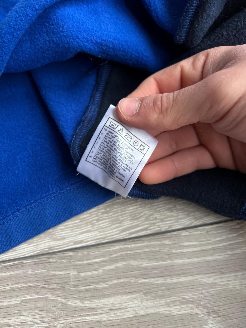 May include: Close-up of a blue and navy blue fleece garment. A white tag with care instructions is visible. The tag includes symbols for washing, ironing, and other care details. The garment appears to be soft and warm.