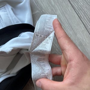 May include: A white garment with a black trim is held by a hand. A tag with text and care symbols is visible. The tag includes washing instructions and fabric composition details.