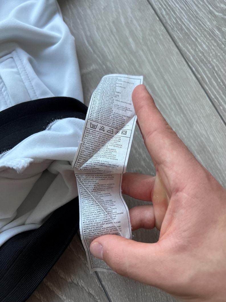 May include: A white garment with a black trim is held by a hand. A tag with text and care symbols is visible. The tag includes washing instructions and fabric composition details.