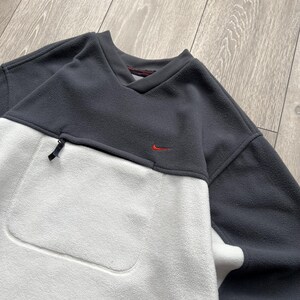 May include: A grey and white fleece pullover with a small red Nike logo. The top has a V-neck, a zipped pocket, and a large front pocket. The fleece material appears soft and warm, ideal for cooler weather.