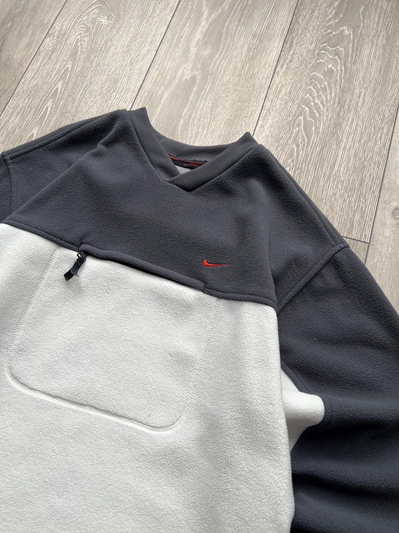 May include: A grey and white fleece pullover with a small red Nike logo. The top has a V-neck, a zipped pocket, and a large front pocket. The fleece material appears soft and warm, ideal for cooler weather.