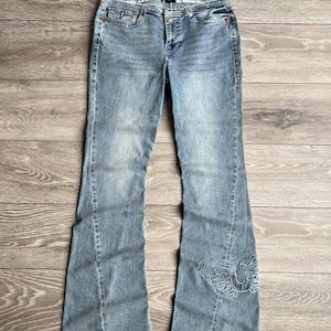 May include: Light blue denim jeans featuring a flared leg silhouette, a classic five-pocket design, a button and zipper fly closure, and a decorative embroidered wing motif in a matching blue thread on the lower portion of the left pant leg.