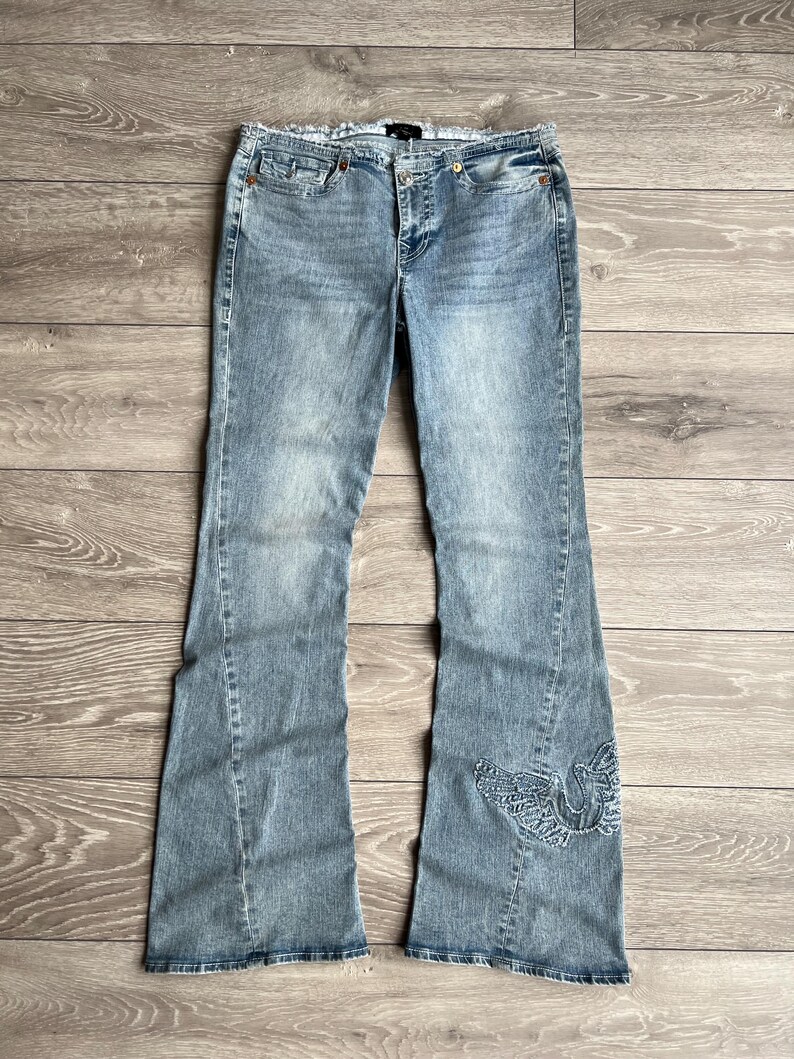 May include: Light blue denim jeans featuring a flared leg silhouette, a classic five-pocket design, a button and zipper fly closure, and a decorative embroidered wing motif in a matching blue thread on the lower portion of the left pant leg.