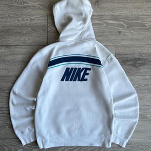 Puede incluir: Sudadera con capucha blanca con la palabra "NIKE" en azul marino en la espalda. La sudadera tiene un dise&ntilde;o de rayas azul marino y turquesa en la parte superior de la espalda. La capucha est&aacute; levantada y las mangas son largas.