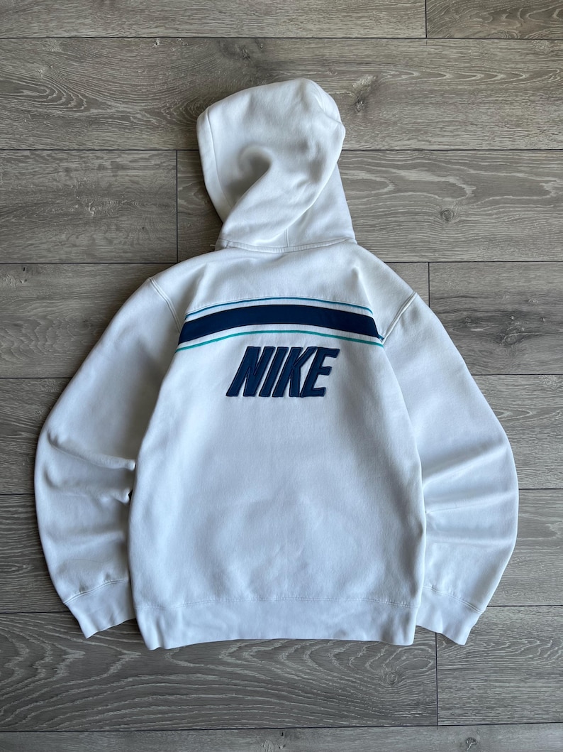 Puede incluir: Sudadera con capucha blanca con la palabra "NIKE" en azul marino en la espalda. La sudadera tiene un dise&ntilde;o de rayas azul marino y turquesa en la parte superior de la espalda. La capucha est&aacute; levantada y las mangas son largas.