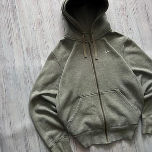 Puede incluir: Una sudadera con capucha gris con cremallera, capucha y bolsillos delanteros. La sudadera tiene un peque&ntilde;o logotipo blanco de Nike en el pecho. La cremallera y los cordones son de un tono gris m&aacute;s oscuro. La sudadera est&aacute; hecha de un material suave y c&oacute;modo.