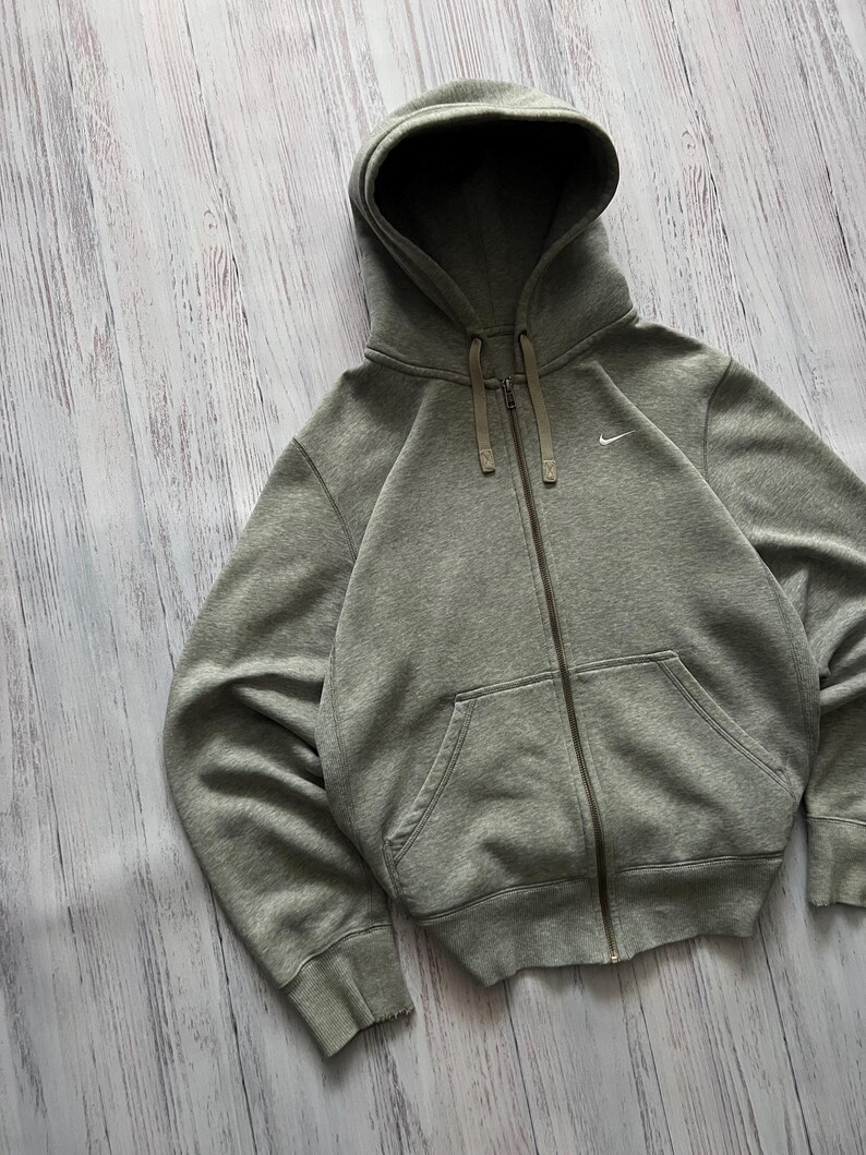 Puede incluir: Una sudadera con capucha gris con cremallera, capucha y bolsillos delanteros. La sudadera tiene un peque&ntilde;o logotipo blanco de Nike en el pecho. La cremallera y los cordones son de un tono gris m&aacute;s oscuro. La sudadera est&aacute; hecha de un material suave y c&oacute;modo.