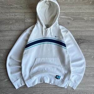 Puede incluir: Sudadera con capucha blanca con bolsillo delantero y dise&ntilde;o de rayas horizontales en azul marino y verde azulado. Presenta el logotipo de Nike swoosh en el pecho y un peque&ntilde;o parche en la parte inferior izquierda.