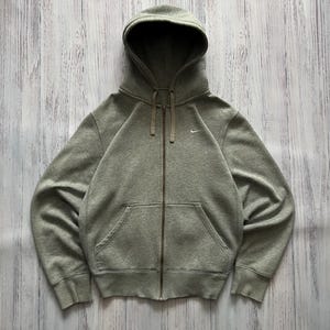 Puede incluir: Una sudadera gris con cremallera, capucha y bolsillos delanteros. El logotipo de Nike es visible en el pecho izquierdo. La sudadera con capucha est&aacute; hecha de un material suave y tiene un cord&oacute;n en la capucha.