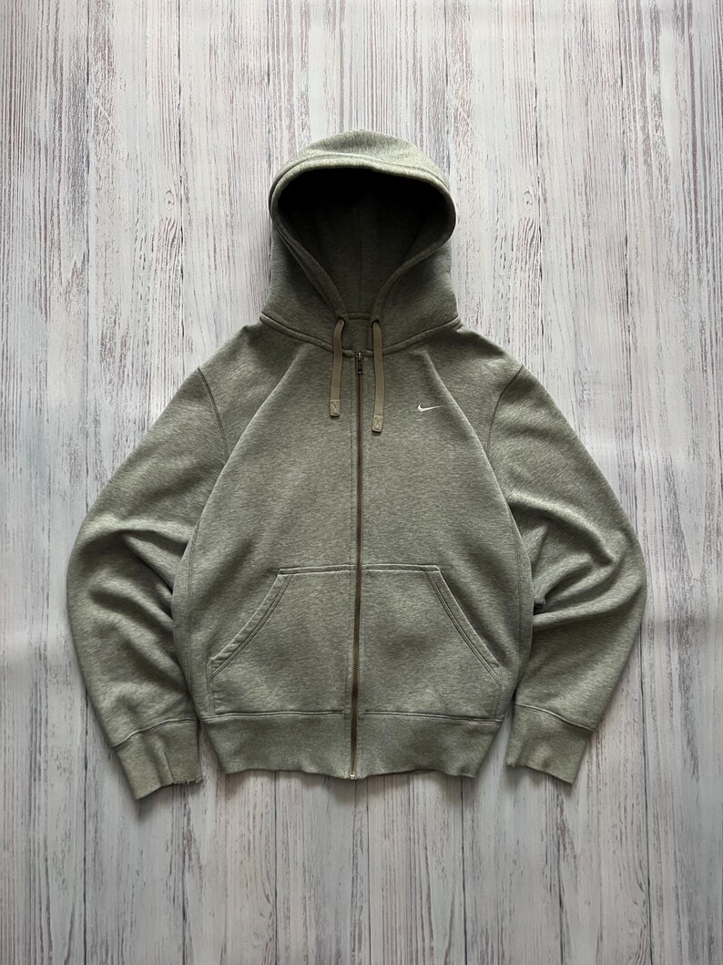 Puede incluir: Una sudadera gris con cremallera, capucha y bolsillos delanteros. El logotipo de Nike es visible en el pecho izquierdo. La sudadera con capucha est&aacute; hecha de un material suave y tiene un cord&oacute;n en la capucha.