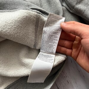Puede incluir: Primer plano del interior de una sudadera gris con forro blanco y afelpado. El exterior de la sudadera es gris liso y se ve una etiqueta blanca con texto negro. La etiqueta proporciona detalles sobre la composici&oacute;n del tejido.