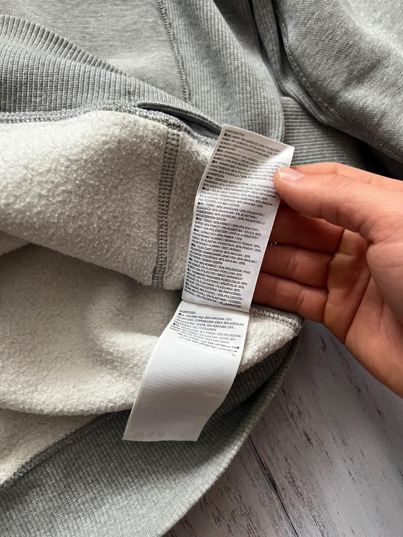 Puede incluir: Primer plano del interior de una sudadera gris con forro blanco y afelpado. El exterior de la sudadera es gris liso y se ve una etiqueta blanca con texto negro. La etiqueta proporciona detalles sobre la composici&oacute;n del tejido.