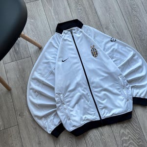 May include: White zip-up jacket with black trim and the Nike logo. Features a Juventus team crest on the chest. The jacket has a black collar and cuffs. The jacket is made of a shiny material.