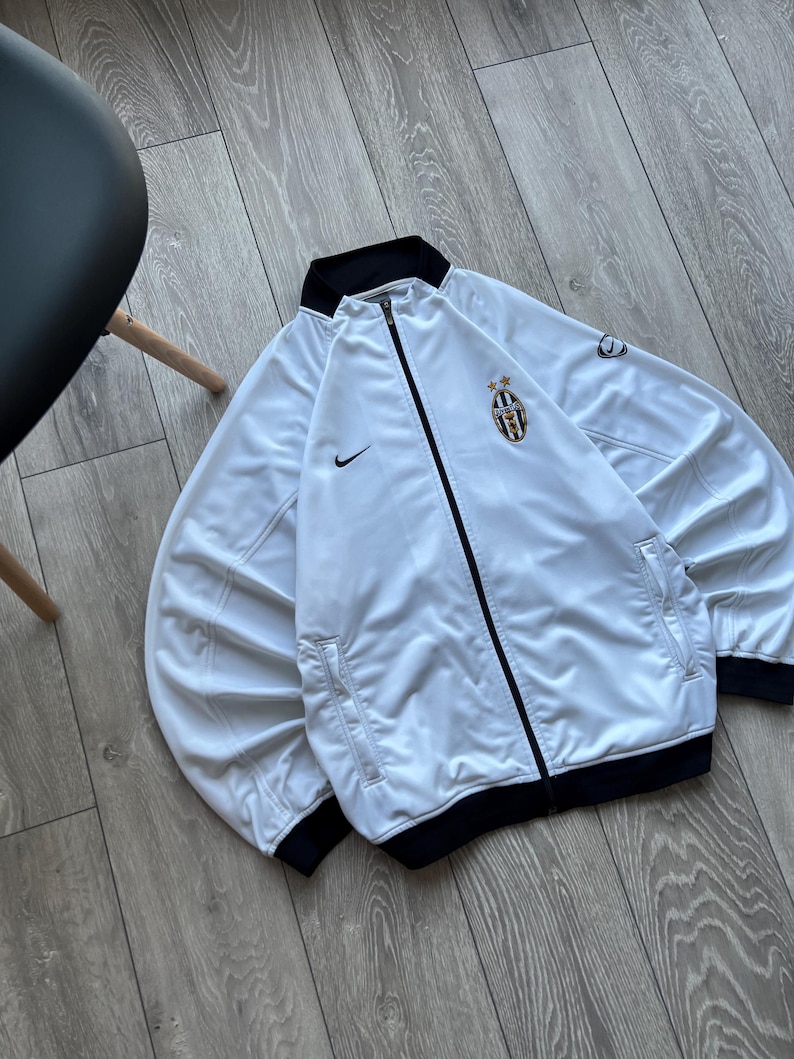May include: White zip-up jacket with black trim and the Nike logo. Features a Juventus team crest on the chest. The jacket has a black collar and cuffs. The jacket is made of a shiny material.
