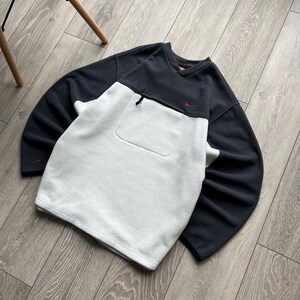 May include: A black and white Nike sweatshirt with a small red logo. The sweatshirt features a front pocket and a zip near the neckline. The sleeves and upper section are black, with a white lower front.