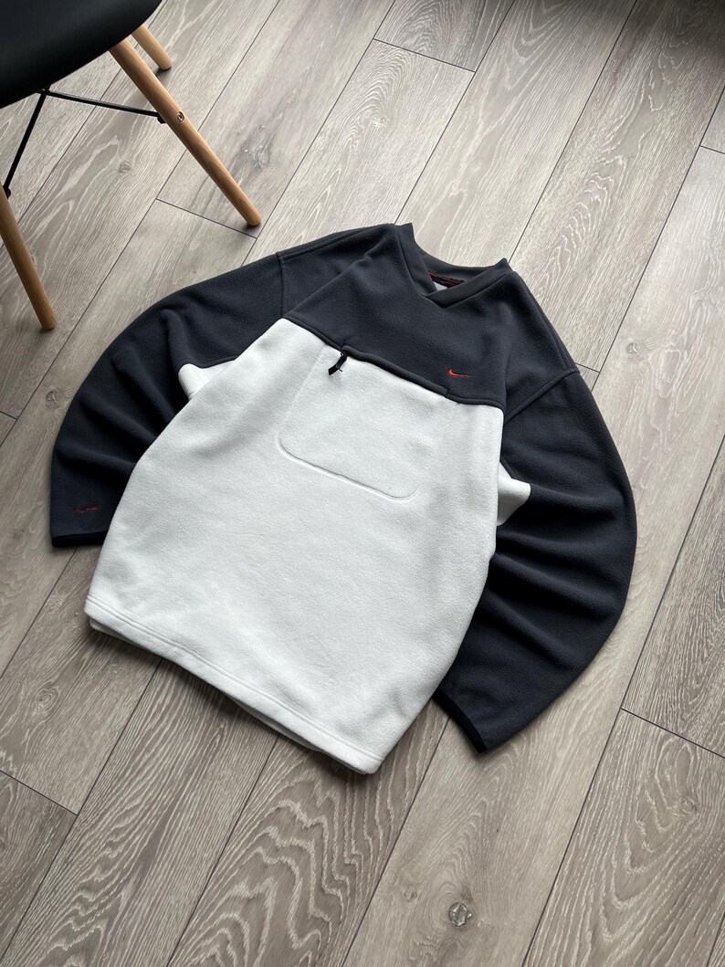 May include: A black and white Nike sweatshirt with a small red logo. The sweatshirt features a front pocket and a zip near the neckline. The sleeves and upper section are black, with a white lower front.