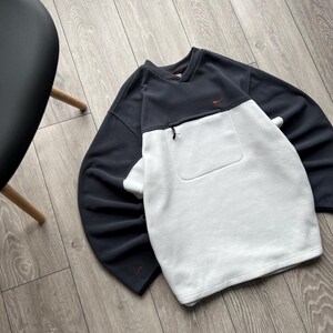 May include: A navy and white fleece sweatshirt. The top half is navy, the bottom half is white. It has a small red logo on the chest and a zipped pocket. The sweatshirt is on a wooden floor.