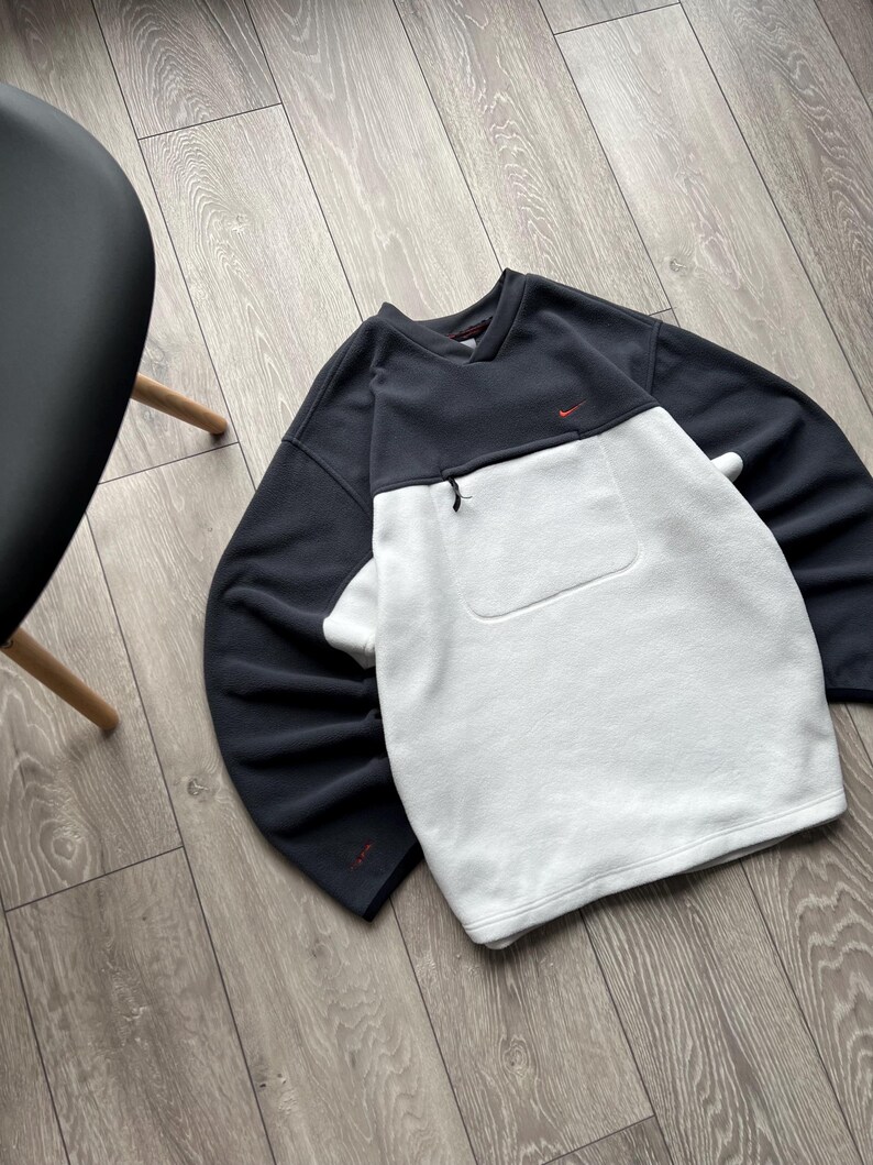 May include: A navy and white fleece sweatshirt. The top half is navy, the bottom half is white. It has a small red logo on the chest and a zipped pocket. The sweatshirt is on a wooden floor.