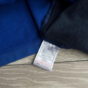 May include: Close-up of a blue and navy blue fleece garment. A white tag with black text is attached, reading "KEEP AWAY FROM FIRE" and "MADE IN MALAYSIA". The garment appears to be made of a soft, warm material.