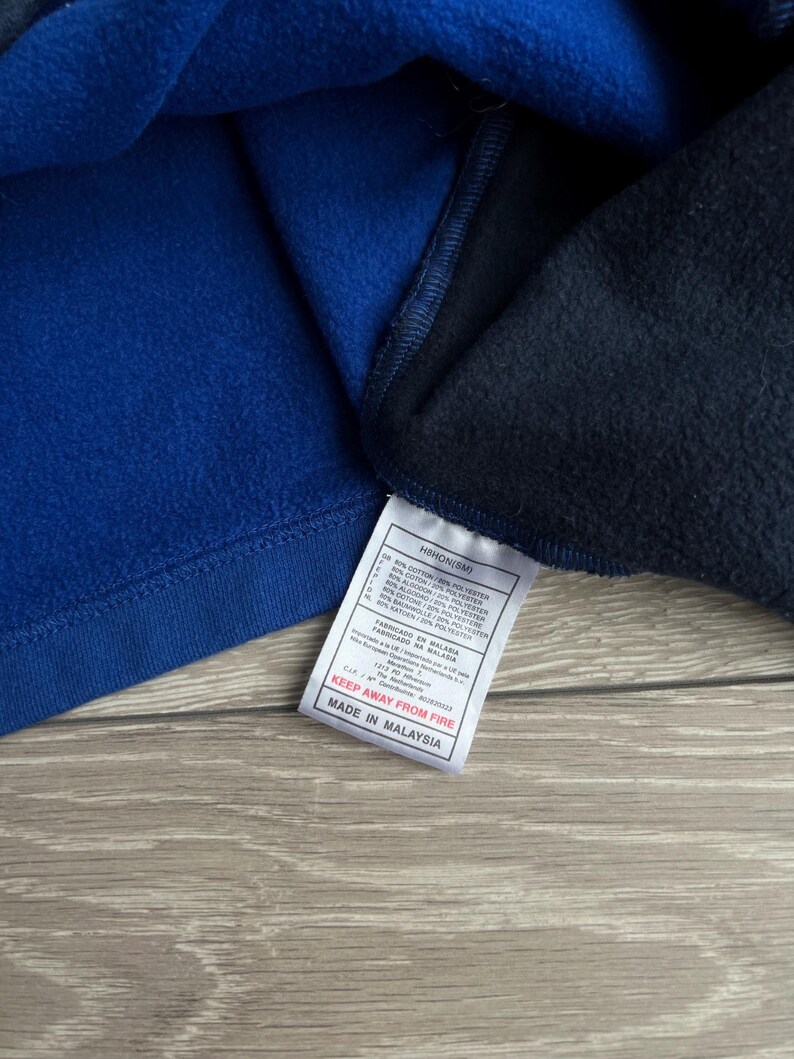 May include: Close-up of a blue and navy blue fleece garment. A white tag with black text is attached, reading "KEEP AWAY FROM FIRE" and "MADE IN MALAYSIA". The garment appears to be made of a soft, warm material.