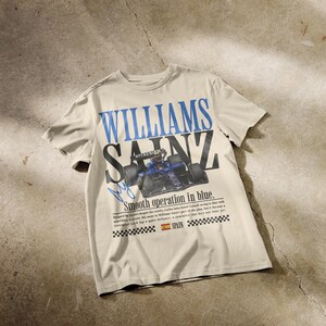 Puede incluir: Camiseta color crema con las palabras "WILLIAMS SAINZ" en azul y negro, y un gr&aacute;fico de coche de carreras. La camiseta incluye el texto "Smooth operation in blue" y una peque&ntilde;a bandera espa&ntilde;ola.