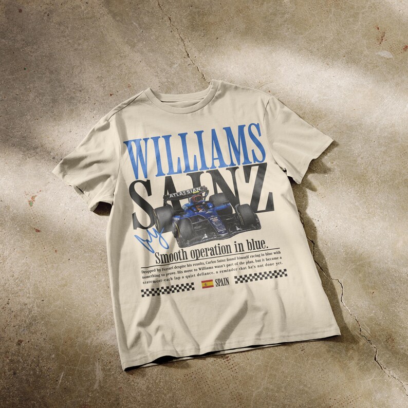 Puede incluir: Camiseta color crema con las palabras "WILLIAMS SAINZ" en azul y negro, y un gr&aacute;fico de coche de carreras. La camiseta incluye el texto "Smooth operation in blue" y una peque&ntilde;a bandera espa&ntilde;ola.