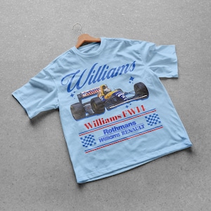 May include: Light blue t-shirt featuring a vintage Williams FW14 Formula 1 race car design. The shirt displays the word "Williams" in a script font, along with the Rothmans and Renault logos. The design includes a checkered flag pattern.