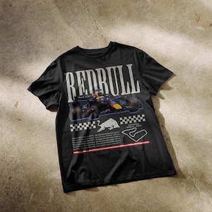 以下が含まれることがあります： 黒いTシャツ。大きな白い文字で「REDRULL」と書かれ、その上にレーシングカーのグラフィックが描かれています。車の真下には白い雄牛のロゴとレーストラックの輪郭があります。デザインには市松模様が含まれています。