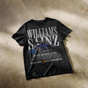 Puede incluir: Camiseta negra con el texto blanco "WILLIAMS SAINZ", con un gr&aacute;fico de coche de carreras azul. La camiseta tambi&eacute;n incluye el texto "Smooth operation in blue" y una peque&ntilde;a bandera espa&ntilde;ola.