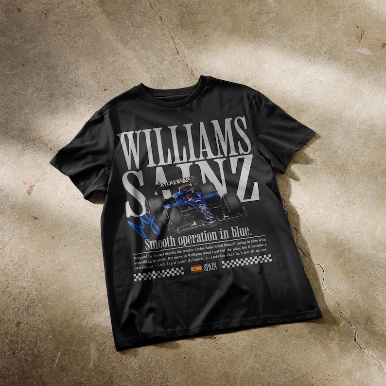 Puede incluir: Camiseta negra con el texto blanco "WILLIAMS SAINZ", con un gr&aacute;fico de coche de carreras azul. La camiseta tambi&eacute;n incluye el texto "Smooth operation in blue" y una peque&ntilde;a bandera espa&ntilde;ola.