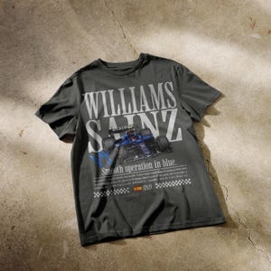 Puede incluir: Camiseta gris oscuro con el texto blanco "WILLIAMS SAINZ" y un gr&aacute;fico de coche de carreras azul. La camiseta tambi&eacute;n incluye el texto "Smooth operation in blue" y la bandera de Espa&ntilde;a.