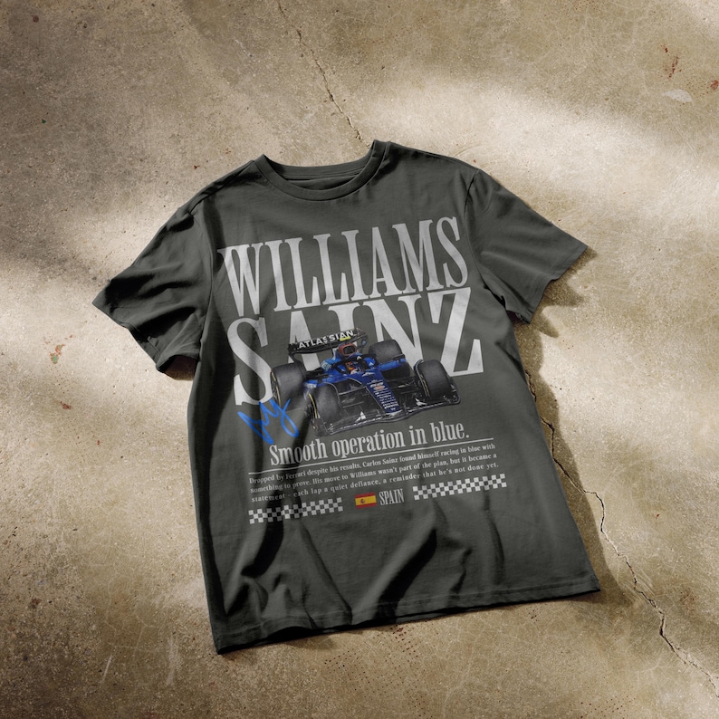 Puede incluir: Camiseta gris oscuro con el texto blanco "WILLIAMS SAINZ" y un gr&aacute;fico de coche de carreras azul. La camiseta tambi&eacute;n incluye el texto "Smooth operation in blue" y la bandera de Espa&ntilde;a.