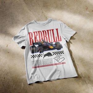 May include: A light grey short-sleeve t-shirt featuring a large red "REDBULL" graphic, a Formula 1 race car, a black bull silhouette, a checkered flag pattern, a track map of the Red Bull Ring in Austria, and small blocks of descriptive text.