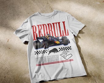 Pode incluir: Uma t-shirt cinzenta clara de manga curta com um grande gráfico vermelho "REDBULL", um carro de corrida de Fórmula 1, a silhueta de um touro preto, um padrão de xadrez, o mapa do circuito Red Bull Ring na Áustria e blocos de texto.