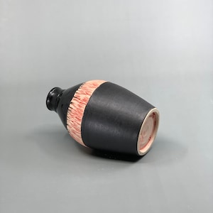 May include: A black ceramic vase with a cylindrical shape and a narrow neck. A band of red and white textured design encircles the middle. The vase is lying on its side against a gray background.