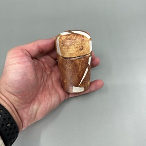 Puede incluir: Un pequeño recipiente de cerámica marrón con un diseño geométrico blanco. El recipiente tiene una tapa y es aproximadamente del tamaño de la mano de una persona.