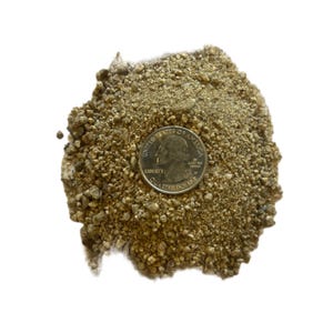 May include: A pile of light brown, granular material with a silver quarter coin on top for size comparison. The quarter shows the profile of George Washington and the words "United States of America". The material appears to be small, irregular pieces.