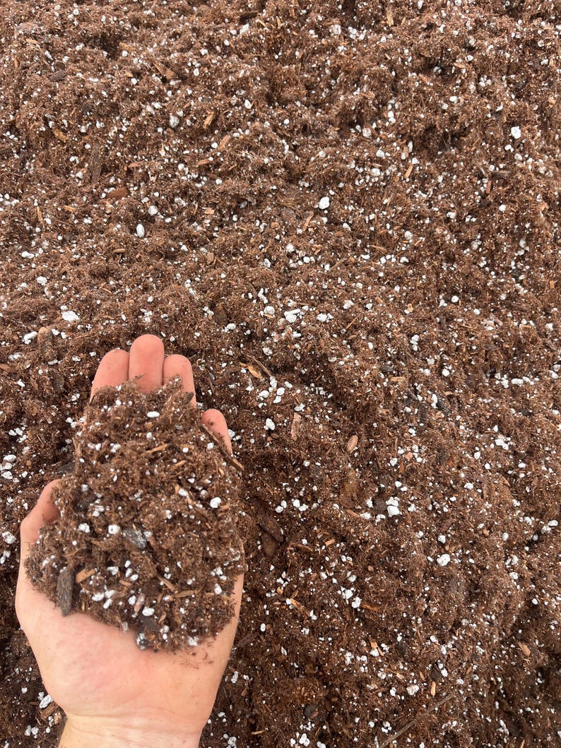 May include: A close-up shot of a handful of brown potting soil with white and light brown flecks. The soil is held in an open hand, with the rest of the soil visible in the background. The soil appears to be a mix of organic matter and other materials.