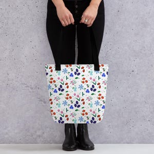 Puede incluir: Bolso tote blanco con un colorido estampado floral con mariquitas rojas, bayas azules y moradas y varias flores. El bolso tiene asas negras. Una persona lleva el bolso, vestida con pantalones y botas negras.
