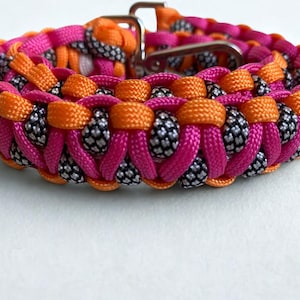 Può includere: Un vivace collare per cani dal design intrecciato, con una combinazione di rosa acceso, arancione e dettagli in bianco e nero. Il collare ha una chiusura in metallo argentato ed è progettato per accessori per animali domestici.