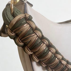 Pode incluir: Close-up de uma bolsa creme com uma alça trançada. A alça é uma combinação de corda verde-oliva e dourada, criando um design texturizado e elegante. Um anel dourado conecta a alça à bolsa.
