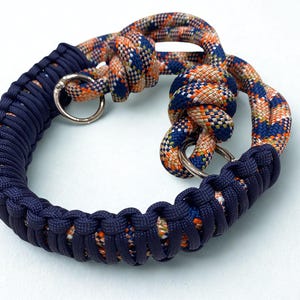 Könnte beinhalten: Ein marineblaues und orangefarbenes Hundehalsband mit silbernen Metallringen. Das Halsband besteht aus geflochtenem Seil und hat ein geflochtenes Design. Das Seil hat ein sich wiederholendes Muster aus Orange, Blau und Weiß.