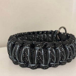 May include: A black and silver paracord bracelet with a metal ring. The bracelet is made of woven black cord with silver accents. The design features vertical silver stripes and a silver ring for attachment.